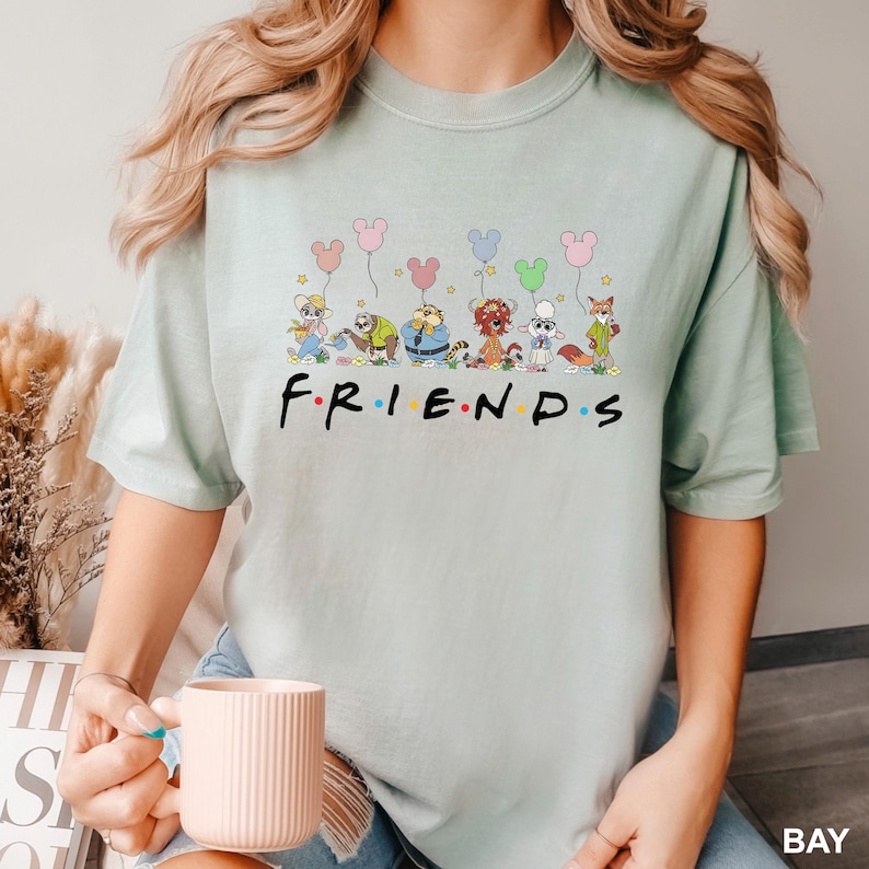 May include: A light green t-shirt featuring the word "FRIENDS" in a colorful font. Above the word are cartoon characters holding Mickey Mouse-shaped balloons. The shirt is a soft, comfortable material.