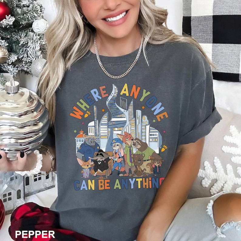 May include: Dark gray t-shirt with the text "WHERE ANYONE CAN BE ANYTHING" in orange, blue, and yellow. The graphic features cartoon animals in front of a city skyline. The shirt has rolled-up sleeves.