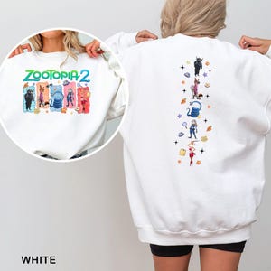 K&ouml;nnte beinhalten: Wei&szlig;es Sweatshirt mit dem Text "ZOOTOPIA 2" in Gr&uuml;n und Blau sowie Zeichentrickfiguren. Die R&uuml;ckseite des Sweatshirts hat ein vertikales Design mit Zeichentrickfiguren und Sternen. Das Wort "WHITE" steht unten.