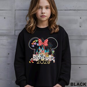 Puede incluir: Sudadera negra con un gr&aacute;fico colorido de un castillo, Mickey Mouse, Minnie Mouse y el texto "Disney Trip 2020". El dise&ntilde;o incluye un lazo rojo con lunares blancos y un arco&iacute;ris de cabezas de Mickey Mouse. La sudadera la lleva una persona joven.