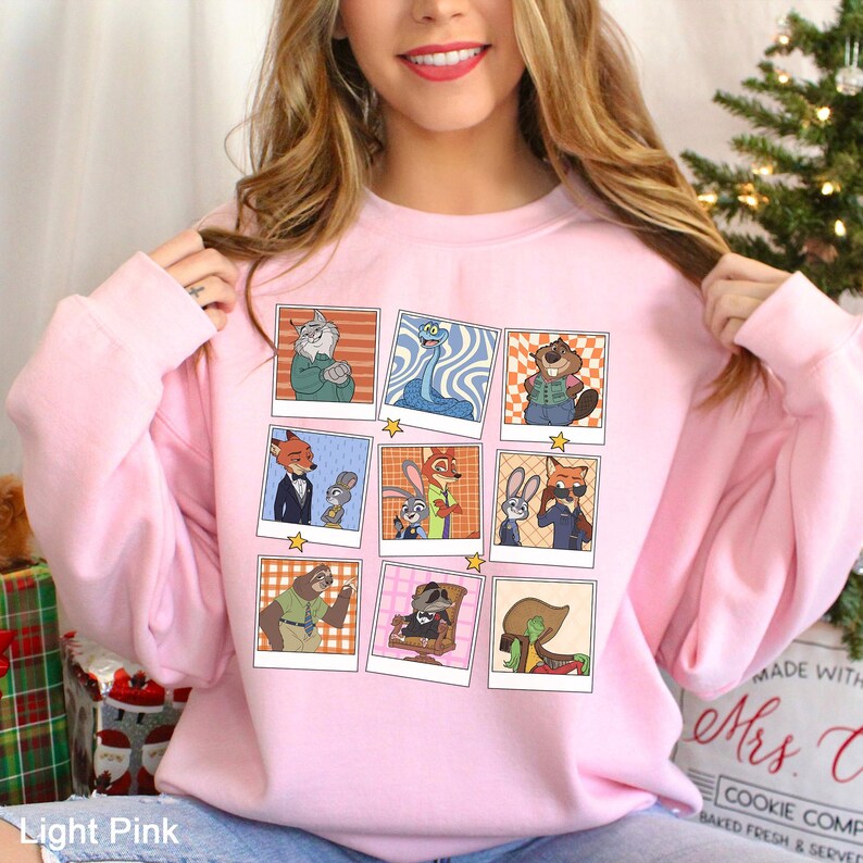 May include: Light pink sweatshirt featuring a grid of cartoon character images in polaroid-style frames. The images include various animal characters in different poses. The sweatshirt is a solid light pink color.