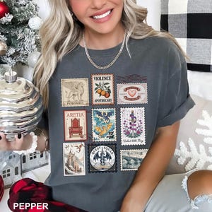 May include: Dark gray t-shirt featuring a collection of postage stamp designs. The stamps depict various illustrations, including a griffin, a throne, and a dragon. The shirt also has the word "PEPPER" printed at the bottom.