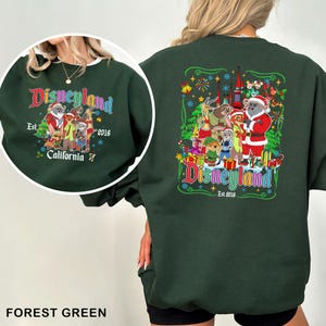 May include: Forest green sweatshirt with a Disneyland graphic. The front features a circular design with text and characters. The back has a larger graphic with the Disneyland logo, characters, and a castle. The sweatshirt is a crew neck.