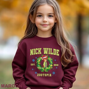 Pu&ograve; includere: Una felpa bordeaux con la scritta "NICK WILDE" in verde, e un'immagine a cartoni animati di Nick Wilde in una ghirlanda natalizia. La felpa include anche il testo "EST. 2016" e "ZOOTOPIA".
