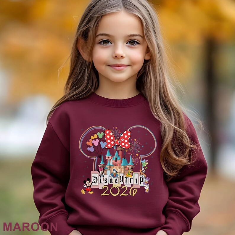 Puede incluir: Sudadera granate con un gr&aacute;fico tem&aacute;tico de Disney. El dise&ntilde;o incluye orejas de Minnie Mouse, un lazo de lunares, un castillo y el texto "Disney Trip 2026". La palabra "MAROON" est&aacute; impresa en la parte inferior.