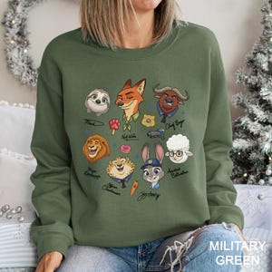 K&ouml;nnte beinhalten: Ein milit&auml;rgr&uuml;nes Sweatshirt mit Cartoon-Illustrationen von Zootopia-Charakteren, darunter Nick Wilde und Judy Hopps. Die Namen der Charaktere sind darunter geschrieben. Der Text "MILITARY GREEN" befindet sich unten.