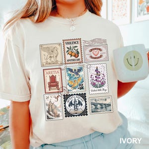 May include: Ivory-colored t-shirt featuring a collection of postage stamp designs. The stamps depict various illustrations, including a griffin, a throne, a dragon, and text such as "VIOLENCE" and "QUEST SQUAD". A white mug with a smiley face is visible.