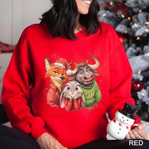 May include: A bright red sweatshirt featuring a graphic of three cartoon characters: a fox wearing a Santa hat, a bull in a green sweater, and a bunny. The word "RED" is printed in white at the bottom right.