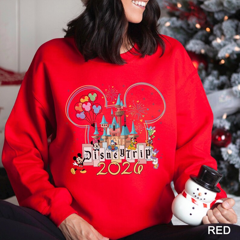 Puede incluir: Una sudadera roja con un dise&ntilde;o de Disney Trip. El dise&ntilde;o incluye un castillo, orejas de Mickey Mouse, globos, fuegos artificiales y varios personajes de Disney. El texto "Disney Trip 2026" tambi&eacute;n se muestra en la sudadera.