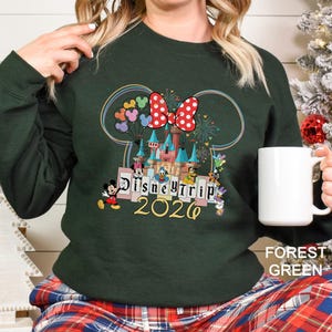 Puede incluir: Sudadera verde bosque con un colorido dise&ntilde;o de Disney Trip 2026. El dise&ntilde;o incluye un castillo, Mickey Mouse, un lazo de Minnie Mouse, globos y otros personajes de Disney. La sudadera es de color liso.