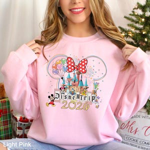 Puede incluir: Sudadera rosa claro con un dise&ntilde;o de Disney Trip 2020. El gr&aacute;fico incluye un castillo, Mickey Mouse, Minnie Mouse, Donald Duck y otros personajes. Tambi&eacute;n presenta un gran lazo rojo con lunares y globos de colores.
