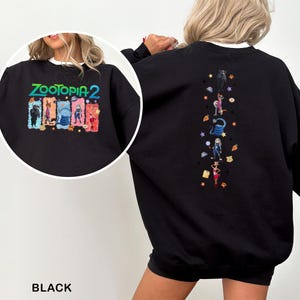 K&ouml;nnte beinhalten: Schwarzes Sweatshirt mit dem Text "ZOOTOPIA 2" in Gr&uuml;n, Gelb und Blau. Das Design auf der Vorderseite zeigt Zeichentrickfiguren. Die R&uuml;ckseite hat ein vertikales Design mit Zeichentrickfiguren und Sternen.