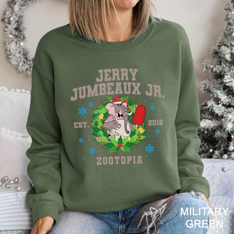 Pu&ograve; includere: Felpa verde militare con la scritta "JERRY JUMBEAUX JR." e un elefante dei cartoni animati con un cappello da Babbo Natale e un ghiacciolo rosso. Il design include una ghirlanda, fiocchi di neve e la parola "ZOOTOPIA".