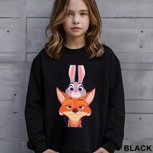 May include: Black sweatshirt featuring a graphic of Judy Hopps the bunny and Nick Wilde the fox from the animated movie Zootopia. The sweatshirt has a crew neck and long sleeves. The word "BLACK" is printed in white at the bottom right.