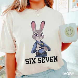 May include: Ivory t-shirt with a cartoon depiction of Judy Hopps from Zootropolis in a police uniform, making the 'OK' hand gesture. The text 'SIX SEVEN' is printed in black below the image. A white mug with a smiley face is also visible.