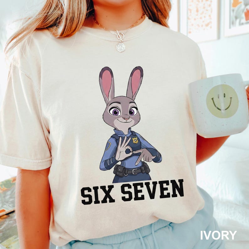 May include: Ivory t-shirt with a cartoon depiction of Judy Hopps from Zootropolis in a police uniform, making the 'OK' hand gesture. The text 'SIX SEVEN' is printed in black below the image. A white mug with a smiley face is also visible.