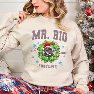 Pu&ograve; includere: Una felpa color sabbia con la scritta "MR. BIG" sopra un personaggio dei cartoni animati con un cappello da Babbo Natale e una ghirlanda. Il design include la scritta "EST. 2016" e "ZOOTOPIA". La felpa ha le maniche lunghe.