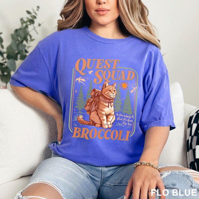 Pode incluir: Uma camiseta azul com um gr&aacute;fico com um gato usando uma mochila, as palavras "QUEST SQUAD" e "BROCCOLI". O design inclui &aacute;rvores, um sol e o texto "No One Wants It, But It's Good For You".
