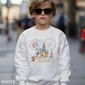 Puede incluir: Sudadera blanca con un colorido gr&aacute;fico del castillo de Disney, Mickey Mouse, globos y el texto "Disney Trip 2020". El dise&ntilde;o est&aacute; dentro de un contorno de oreja de Mickey Mouse. La sudadera la lleva una persona con gafas de sol.