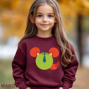 May include: Maroon sweatshirt featuring a cartoon mouse design with a green face, orange ears, and a blue tie. The word "MAROON" is printed at the bottom. The sweatshirt is worn by a young girl with long brown hair.