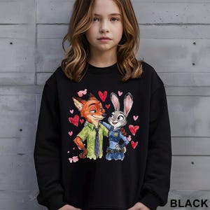 Pu&ograve; includere: Felpa nera con un'illustrazione ad acquerello di Nick Wilde e Judy Hopps di Zootropolis, circondati da cuori rossi. La parola "BLACK" &egrave; stampata in basso a destra.