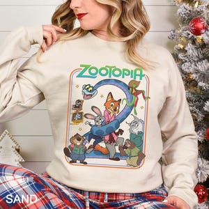 May include: A sand-colored sweatshirt featuring a colorful graphic of characters from the animated film "Zootopia." The design includes the film's title in green, along with images of a fox, rabbit, and other characters. The sweatshirt has long sleeves.