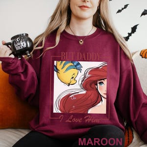 May include: Maroon sweatshirt with a graphic of Ariel and Flounder from The Little Mermaid, and the text "BUT DADDY - I Love Him." The word "MAROON" is printed at the bottom. The sweatshirt is a solid maroon colour.
