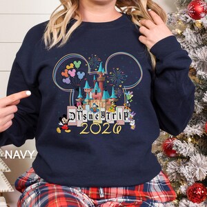 Puede incluir: Sudadera azul marino con un dise&ntilde;o colorido del castillo de Disney, fuegos artificiales y globos. El texto "Disney Trip 2026" se muestra debajo, con Mickey Mouse y otros personajes. La palabra "NAVY" est&aacute; impresa en la parte inferior izquierda.
