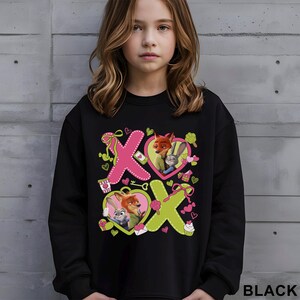 Peut inclure: Sweat-shirt noir avec un motif graphique des personnages de Zootopie, Nick Wilde et Judy Hopps. Le motif comprend des c&oelig;urs roses et verts, les lettres "X" et "O" et d'autres &eacute;l&eacute;ments d&eacute;coratifs. Le mot "BLACK" est imprim&eacute; en bas &agrave; droite.