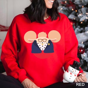 May include: A red sweatshirt featuring a cartoon illustration of a woman wearing glasses, a navy blue blazer, and a polka-dot shirt. The design includes large, tan mouse-ear-like shapes on either side of the woman's head. The word "RED" is printed in the bottom right corner.