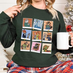 May include: Forest green sweatshirt featuring a grid of cartoon character polaroid-style images. The images include various anthropomorphic animals in different outfits and poses. A white mug is held in the right hand.
