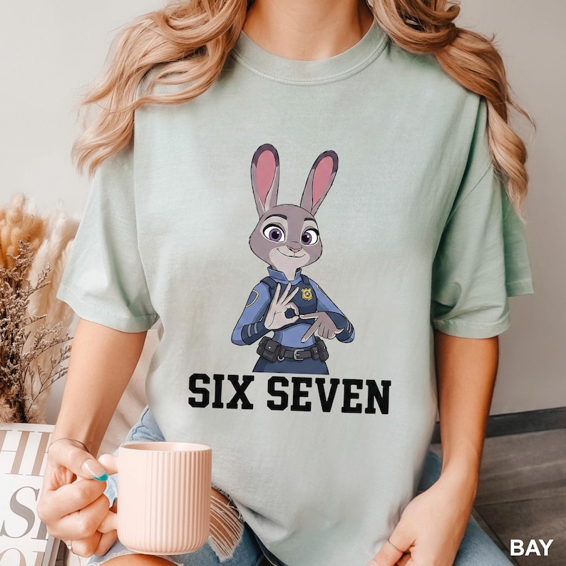 May include: A pale green t-shirt with a cartoon illustration of Judy Hopps from Zootropolis, in a police uniform and making a hand gesture. The words "SIX SEVEN" are printed in bold, black letters below the image.