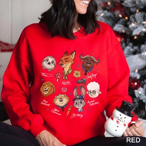 K&ouml;nnte beinhalten: Ein rotes Sweatshirt mit Cartoon-Illustrationen von Zootopia-Charakteren, darunter Flash, Nick Wilde, Judy Hopps und Chief Bogo. Das Design enth&auml;lt Charakternamen und eine kleine Schneemannfigur.