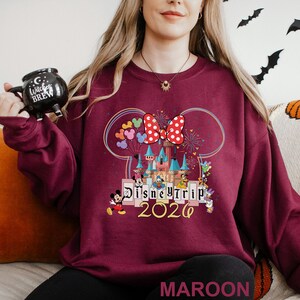Puede incluir: Sudadera granate con un dise&ntilde;o de Disney Trip 2020, con Mickey Mouse, Minnie Mouse y otros personajes. El dise&ntilde;o incluye un castillo, globos y fuegos artificiales. La palabra "MAROON" est&aacute; impresa en la parte inferior.