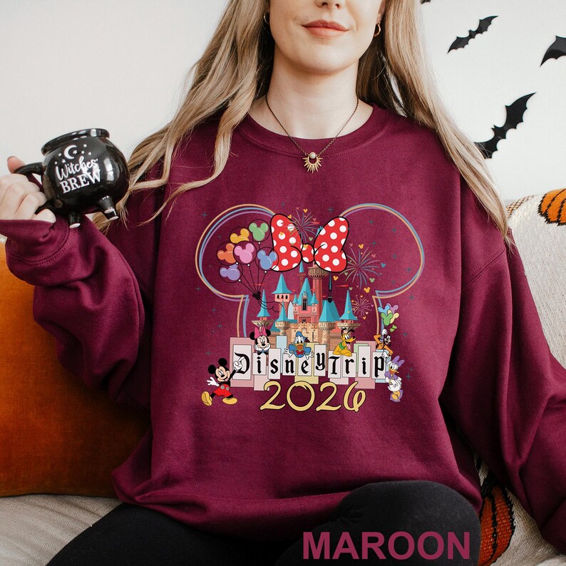 Puede incluir: Sudadera granate con un dise&ntilde;o de Disney Trip 2020, con Mickey Mouse, Minnie Mouse y otros personajes. El dise&ntilde;o incluye un castillo, globos y fuegos artificiales. La palabra "MAROON" est&aacute; impresa en la parte inferior.