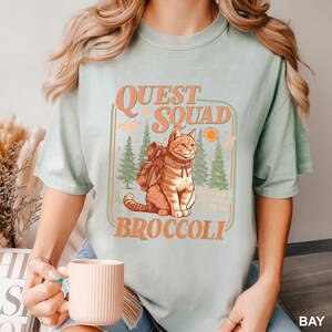 Pode incluir: Camiseta verde claro com um gr&aacute;fico de um gato com uma mochila, as palavras "Quest Squad Broccoli" e uma cena de floresta em laranja e verde. A camiseta &eacute; de estilo casual.