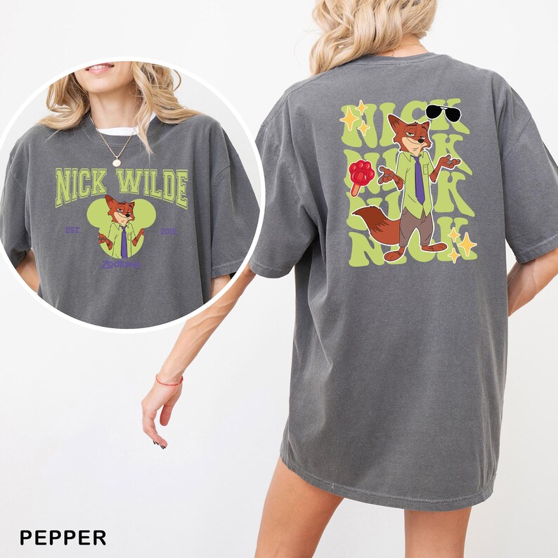 Op de afbeelding: Een antracietgrijs t-shirt met een afbeelding van het personage Nick Wilde. De voorkant toont de naam Nick Wilde boven een Mickey-vormig icoon. De achterkant toont een grote illustratie van het personage met een zonnebril, een ijsje en tekst.
