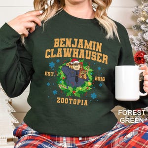 Pu&ograve; includere: Felpa verde foresta con Benjamin Clawhauser di Zootropolis. Il design include il testo "BENJAMIN CLAWHAUSER" sopra un'immagine di Clawhauser con un cappello da Babbo Natale, circondato da una ghirlanda, con il testo "EST. 2016 Zootopia".
