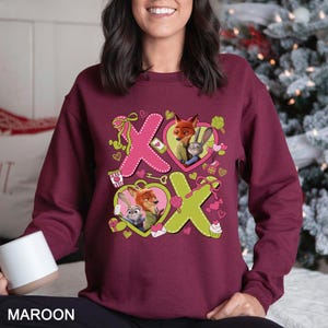 Peut inclure: Sweat-shirt bordeaux avec les personnages Nick Wilde et Judy Hopps de Zootopie. Le motif comprend des formes X et O roses et vertes avec des cadres en forme de c&oelig;ur, des cupcakes et des bo&icirc;tes cadeaux. Le mot "MAROON" est imprim&eacute; en bas.