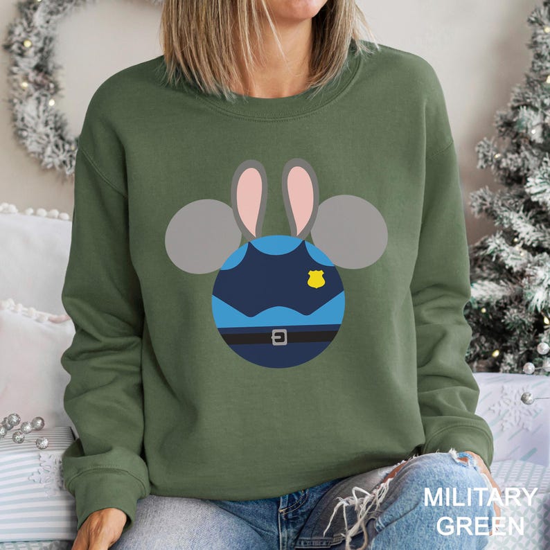 May include: Military green sweatshirt featuring a cartoon design of a police officer bunny. The bunny's head is a navy blue circle with a belt, badge, and gray ears with pink insides. The text "MILITARY GREEN" is at the bottom.