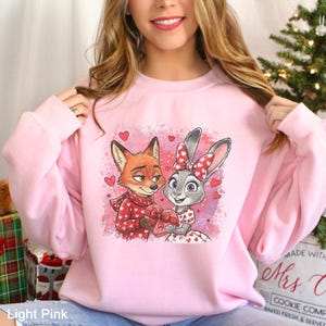 May include: Light pink sweatshirt featuring a cartoon fox and bunny holding a heart-shaped gift. The fox wears a red scarf and the bunny has a bow. Small hearts surround the characters. The text "Light Pink" is at the bottom.