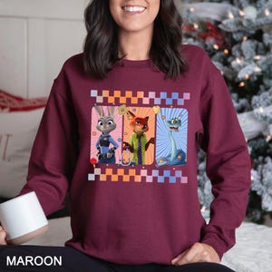 May include: Maroon sweatshirt featuring three cartoon characters in rectangular frames. The characters include a rabbit in a police uniform, a fox in a green suit, and a blue snake. The design is framed with a checkered pattern in orange, white, and blue.