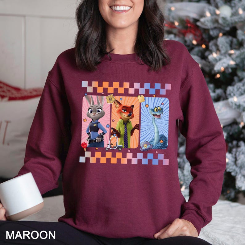 May include: Maroon sweatshirt featuring three cartoon characters in rectangular frames. The characters include a rabbit in a police uniform, a fox in a green suit, and a blue snake. The design is framed with a checkered pattern in orange, white, and blue.