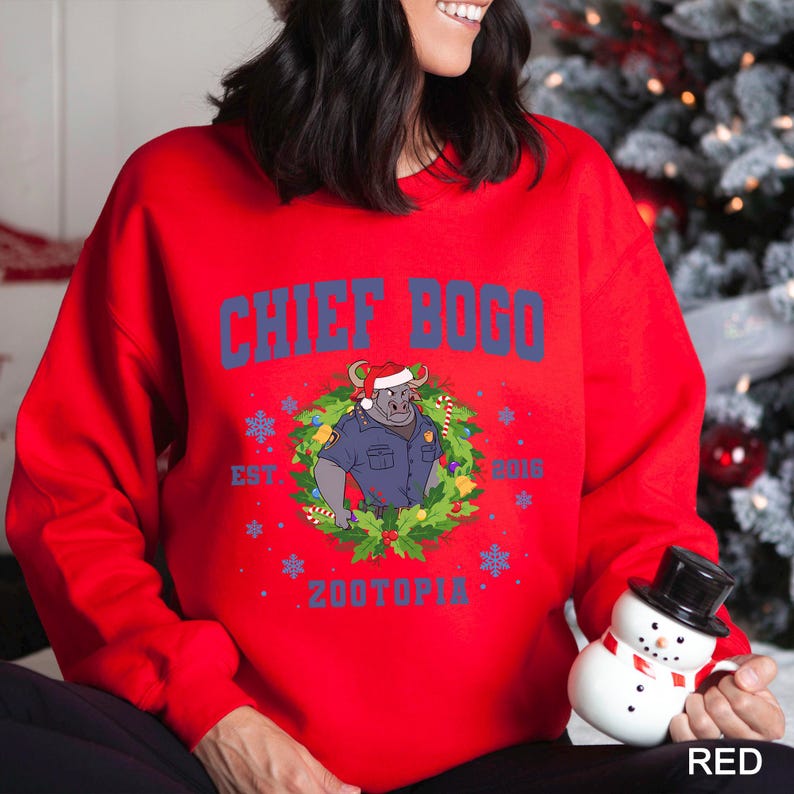 Pu&ograve; includere: Una felpa rossa con la scritta "CHIEF BOGO" in blu, con un'illustrazione di un toro in uniforme da poliziotto con un cappello di Babbo Natale. Il design include una ghirlanda, fiocchi di neve e la parola "ZOOTOPIA".