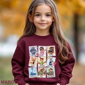 Pu&ograve; includere: Felpa bordeaux con ritratti di personaggi dei cartoni animati. Il design include Chief Bogo, Judith, Mr. Big, Finnick, Nick Wilde, Judy Hopps, Garg de snack, Flash e Nibbles Maplastick. La parola "MAROON" &egrave; stampata in basso.