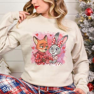May include: A sand-colored sweatshirt featuring a cartoon fox and bunny holding a heart-shaped gift. The fox wears a red scarf and sweater with heart patterns. The bunny wears a dress and bow with heart patterns. Red hearts are scattered around the characters.