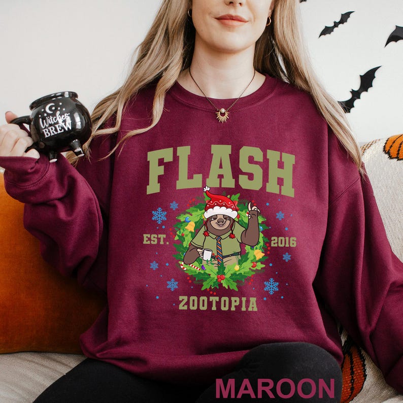 Pu&ograve; includere: Felpa bordeaux con la scritta "FLASH" in lettere verdi. Sotto, un bradipo dei cartoni animati con un cappello da Babbo Natale e una ghirlanda. Anche il testo "EST. 2016 ZOOTOPIA" &egrave; sulla felpa. Una tazza nera con la scritta "Witches Brew" &egrave; tenuta nella mano sinistra.