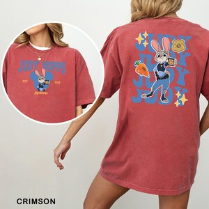 Op de afbeelding: Een karmozijnrood t-shirt met korte mouwen en een afbeelding van Judy Hopps uit Zootopia. De achterkant toont een grote blauwe golvende tekst met Judy Hopps, een wortel en een politiebadge. De voorkant heeft een kleinere Judy Hopps afbeelding.
