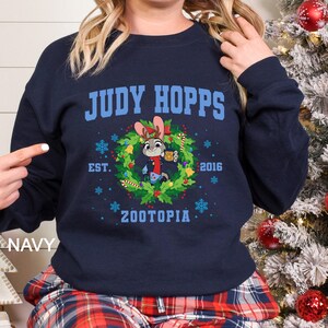 Pu&ograve; includere: Felpa blu navy con la scritta "JUDY HOPPS" sopra un disegno a ghirlanda con il personaggio Judy Hopps di Zootropolis. La ghirlanda include fiocchi di neve, agrifoglio e la scritta "EST. 2016 ZOOTOPIA".