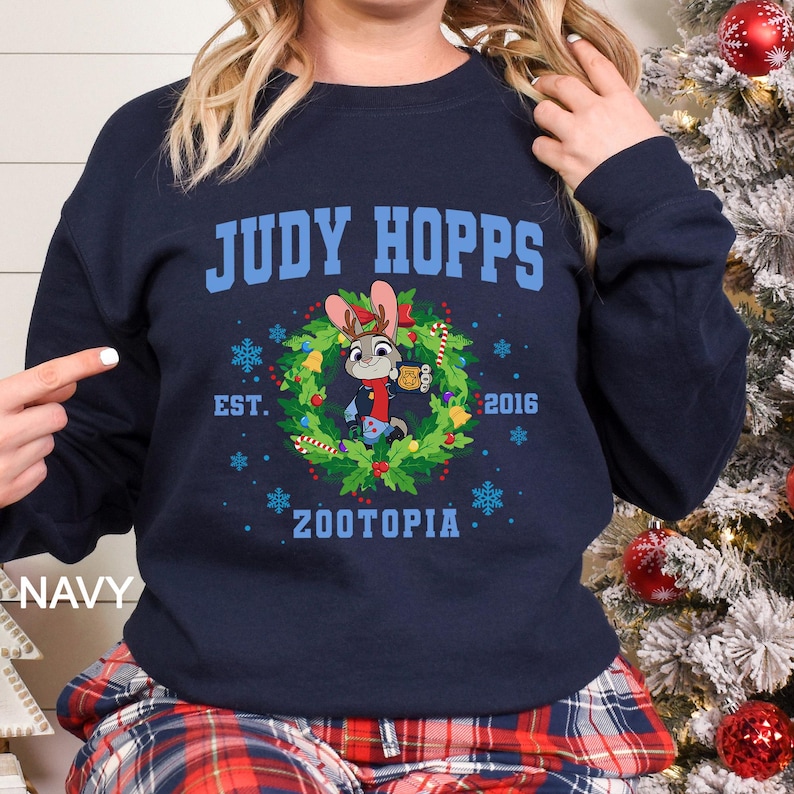 Pu&ograve; includere: Felpa blu navy con la scritta "JUDY HOPPS" sopra un disegno a ghirlanda con il personaggio Judy Hopps di Zootropolis. La ghirlanda include fiocchi di neve, agrifoglio e la scritta "EST. 2016 ZOOTOPIA".